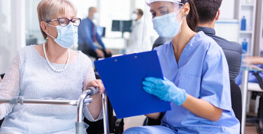 Disabled senior woman having a discussion with medical specialist about treatment in hospital hallway wearing face mask against coronavirus. Patient and medical staff in waiting area.